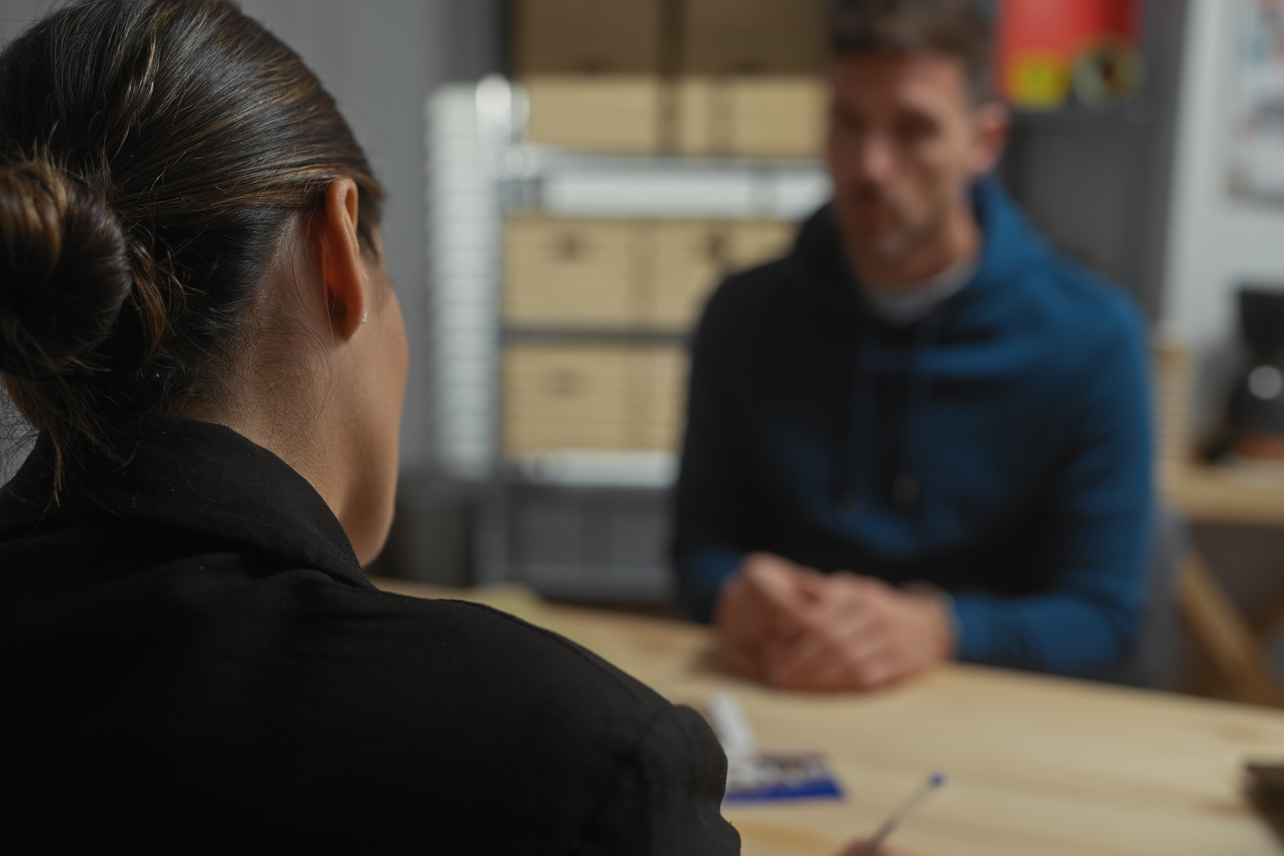 Close-up of a person with a bun facing a blurred man in a blue jacket at a table, suggesting an interview or meeting.