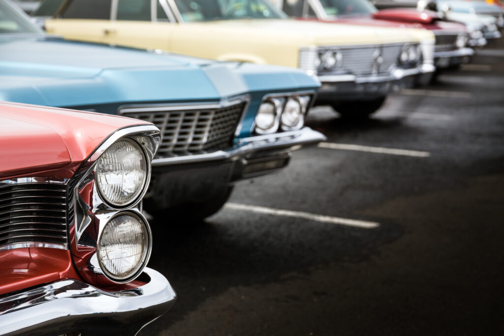 Lineup of vintage American cars parked in a row, focusing on the chrome grilles and dual headlights of the nearest red car