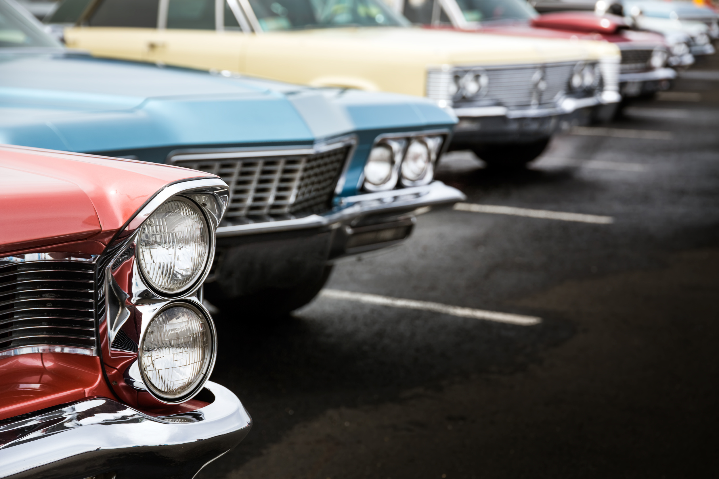 Lineup of vintage American cars parked in a row, focusing on the chrome grilles and dual headlights of the nearest red car