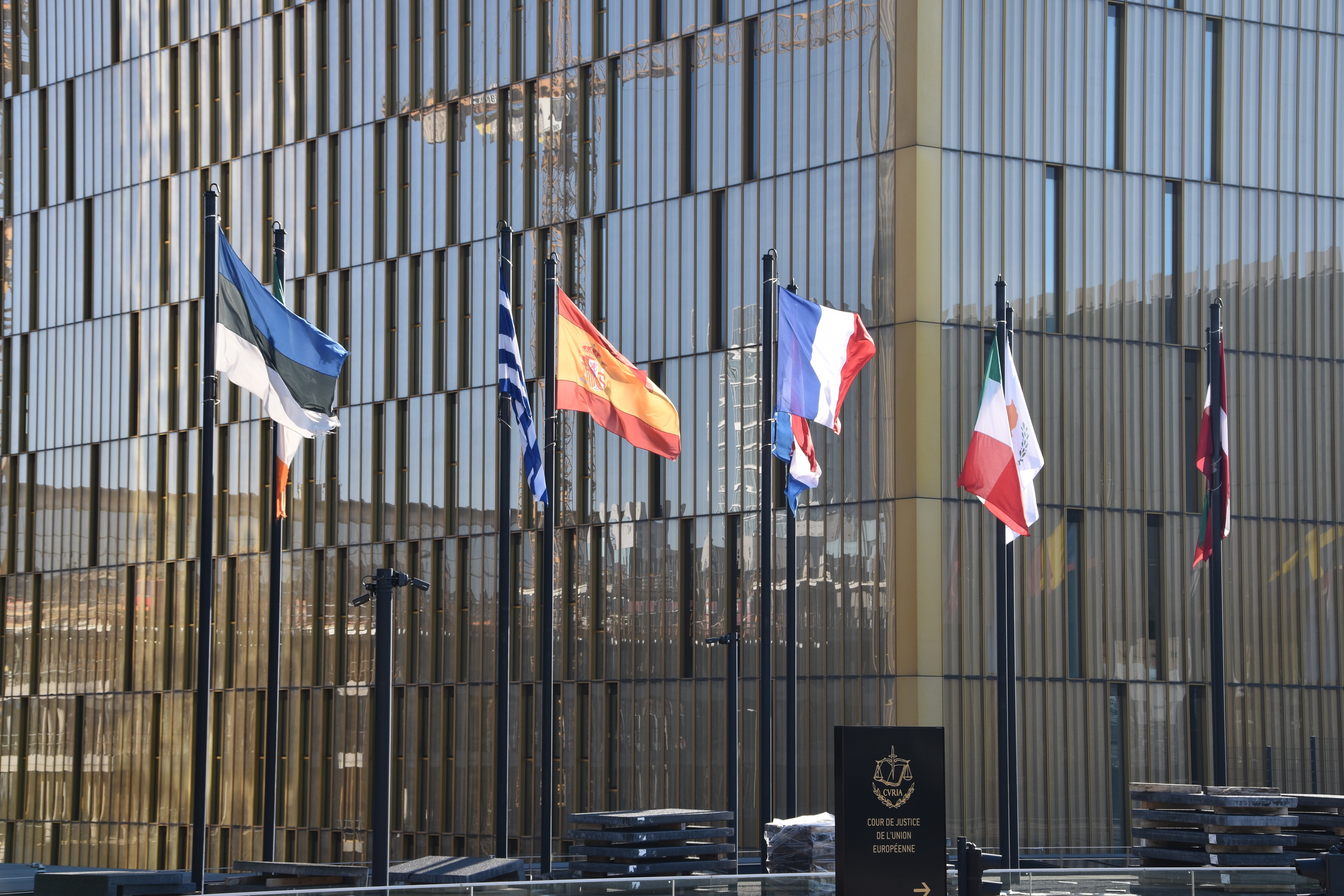 Flags of multiple EU countries line flagpoles in front of a tall glass office building with reflective windows.