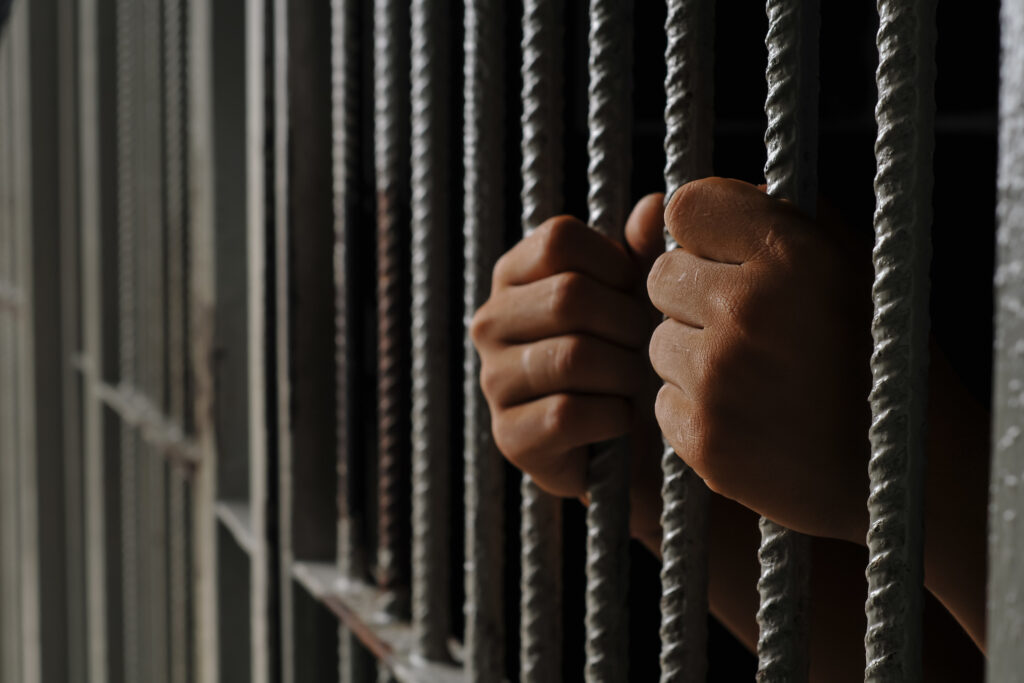 Close up of a prisoner hand gripping rusty iron bars behind a jail cell or detention center.
